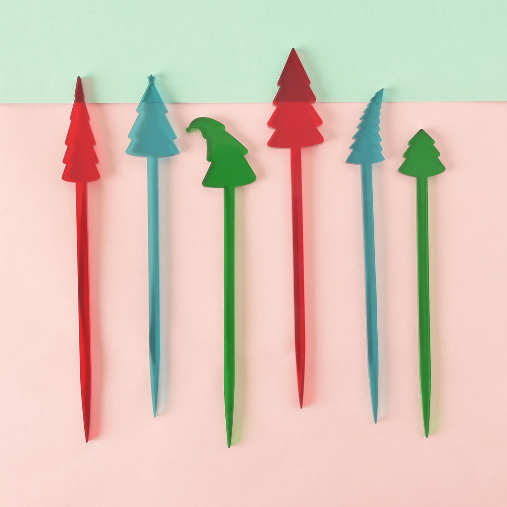 A set of six unique Christmas tree-shaped cocktail stirrers in red, green, and blue. Each stirrer features a slightly different tree shape, including one green tree with a curved, pointed top like a Santa hat. They are arranged side-by-side on a background of solid pink and mint green.

