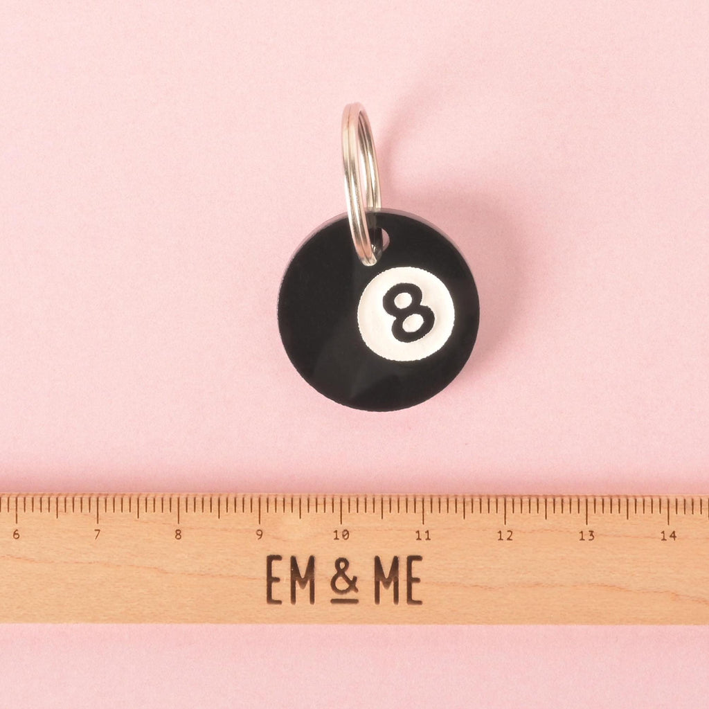Black acrylic 8-ball pet ID tag with a silver keyring, shown on a pink background above a ruler for size reference. Perfect for dogs and cats.