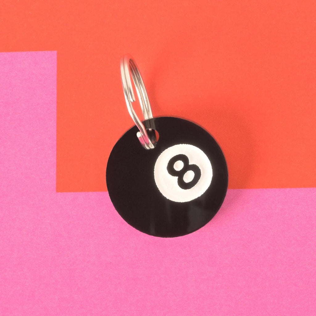 Round black acrylic pet ID tag shaped like an 8-ball with silver keyring, displayed on a pink and orange geometric background.