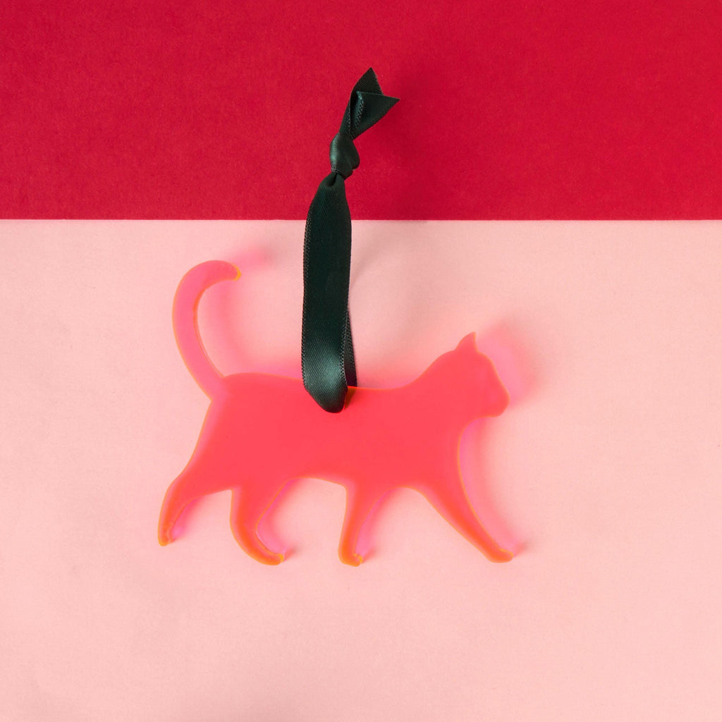 A laser-cut cat silhouette ornament made from vibrant translucent fluorescent pink acrylic, featuring a long tail and hanging from a dark green satin ribbon. The ornament is displayed against a two-tone pink and red background.