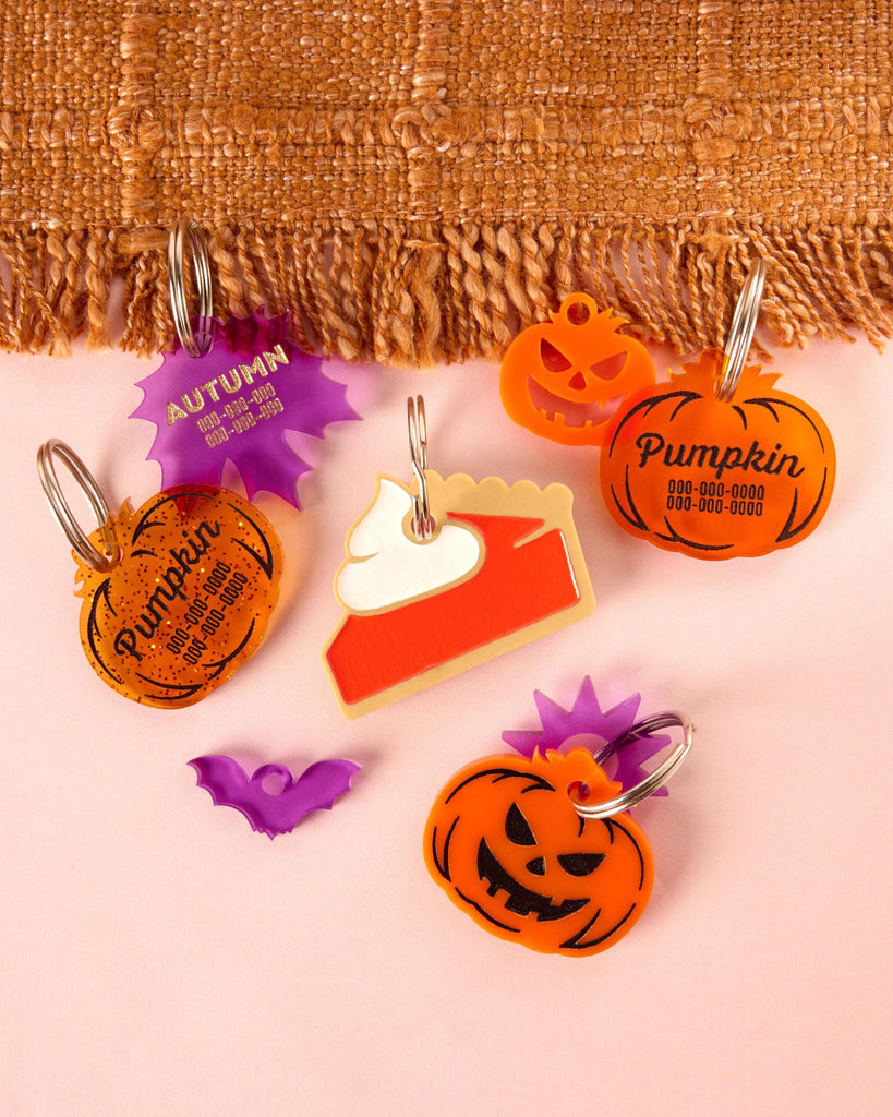 A collection of custom, personalized acrylic pet tags and charms: three distinct pumpkin tags (glitter, plain orange, and jack-o'-lantern), a layered pie slice tag with whipped cream, and a purple starburst tag for 'Autumn'. Also visible is a mini purple bat charm.