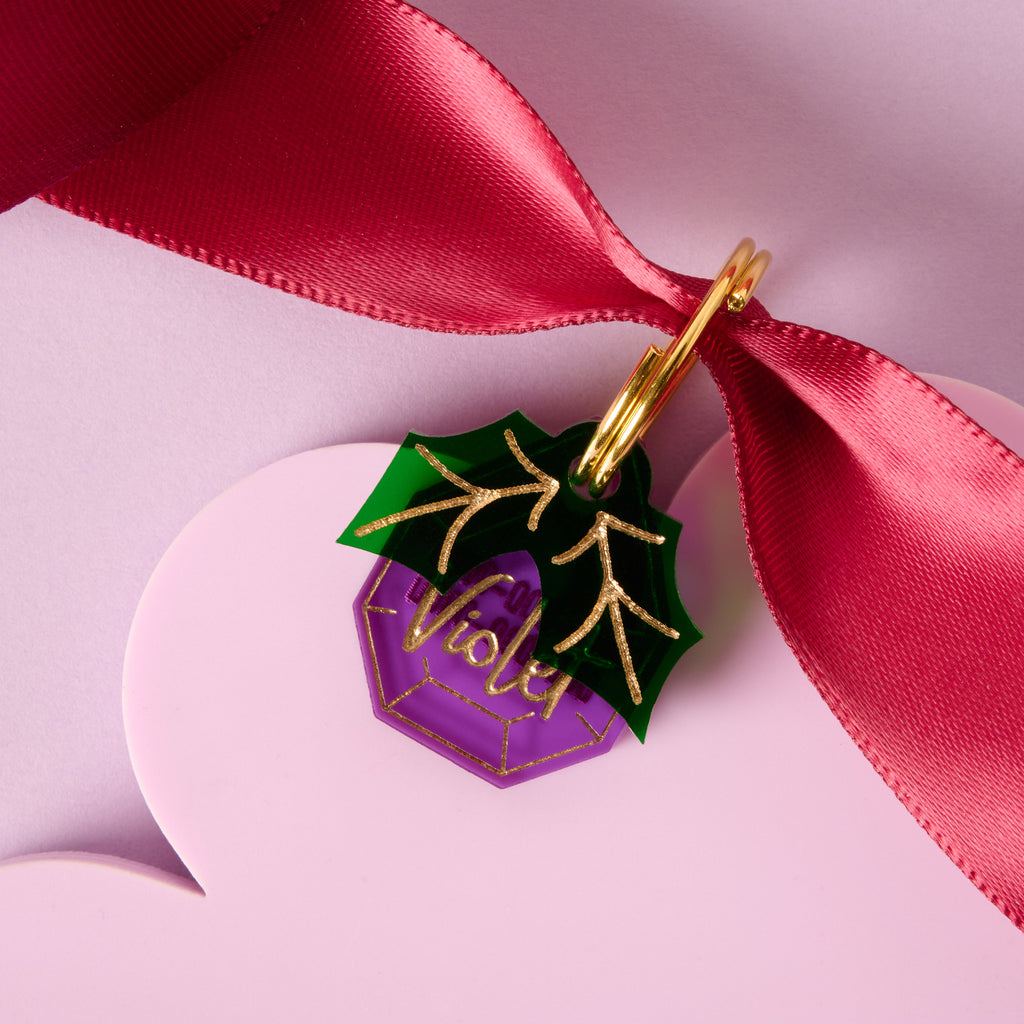Close-up of a purple faceted acrylic pet ID tag, personalized with 'Violet' in gold script. A green acrylic holly leaf charm with gold lines is layered on top. The tag is attached to a gold ring and displayed on a red ribbon against a purple and pink background.