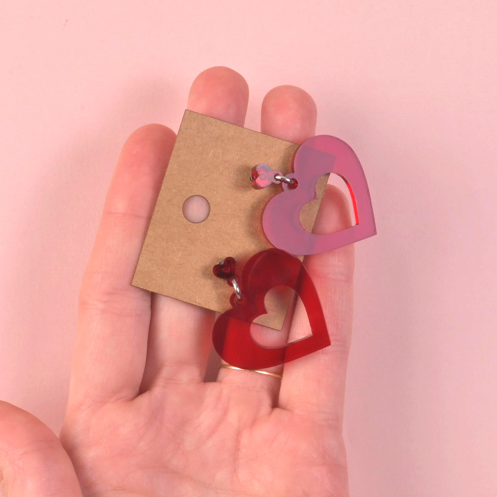 A hand holds two acrylic heart-shaped cutout earrings—one opaque red and one transparent pink—along with a craft paper earring card, illustrating the size and available color options.