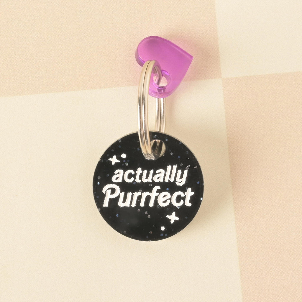 A close-up product photo of a round, black glittery pet tag with a purple heart charm attached to the top. The tag is engraved with the words "actually Purrfect" and is shown with a silver split key ring on a checkered pink and white background.