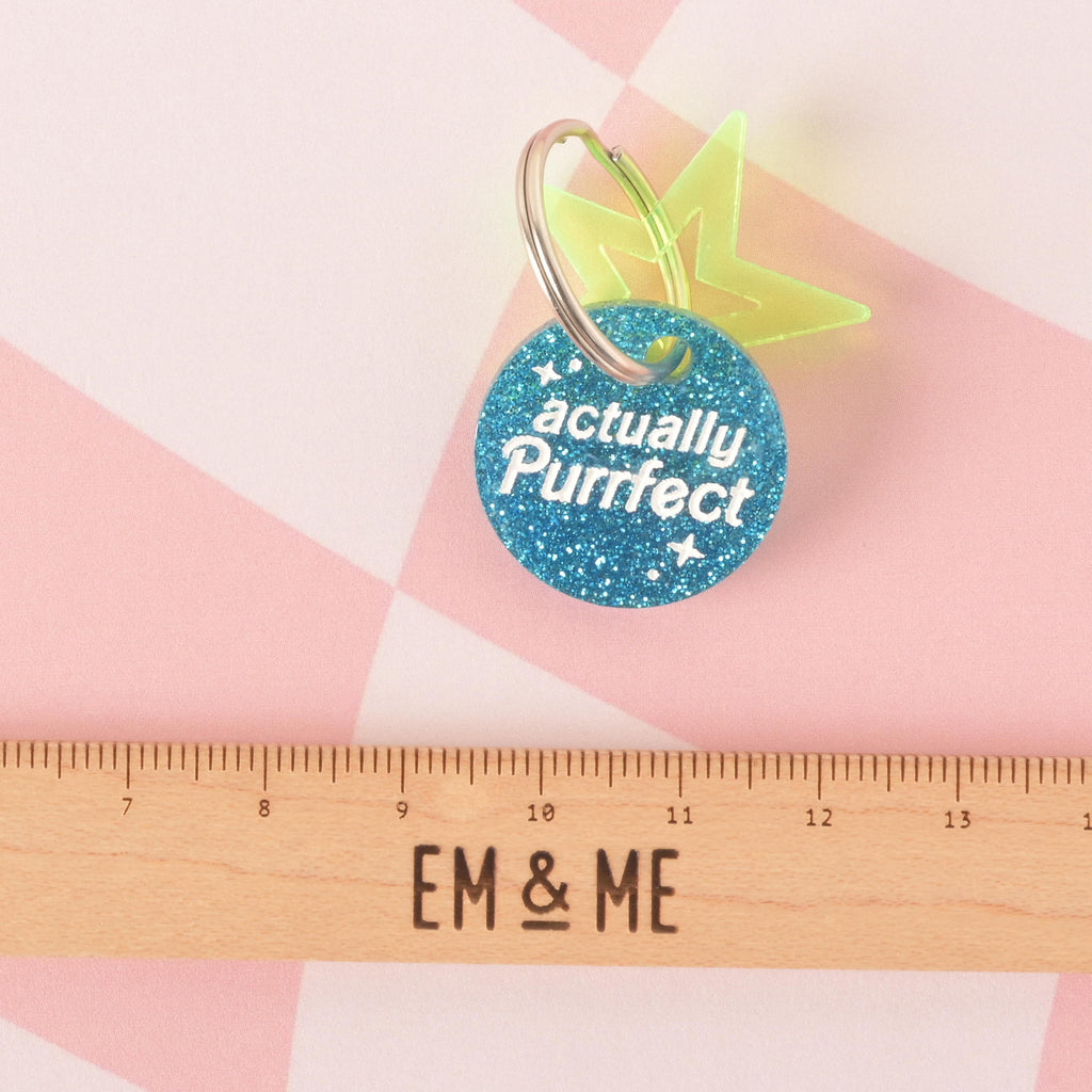 A close-up product photo of a round, blue glittery pet tag with a neon green star charm attached to the top. The tag is engraved with the words "actually Purrfect" and is shown with a silver split key ring. A wooden ruler with the words "EM & ME" is positioned below the tag to show its size.