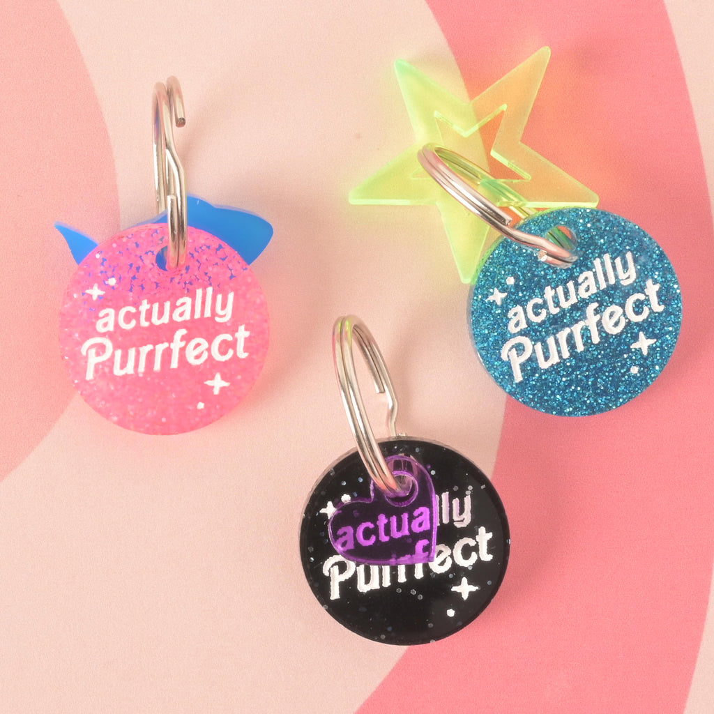 A product photo showing three round, glittery pet ID tags on a checkered pink and white background. The tag on the top left is pink with a blue cat ear shape. The tag on the top right is blue with a neon green star. The tag at the bottom is black with a purple heart. All three tags are engraved with the words "actually Purrfect" and have a silver split key ring.