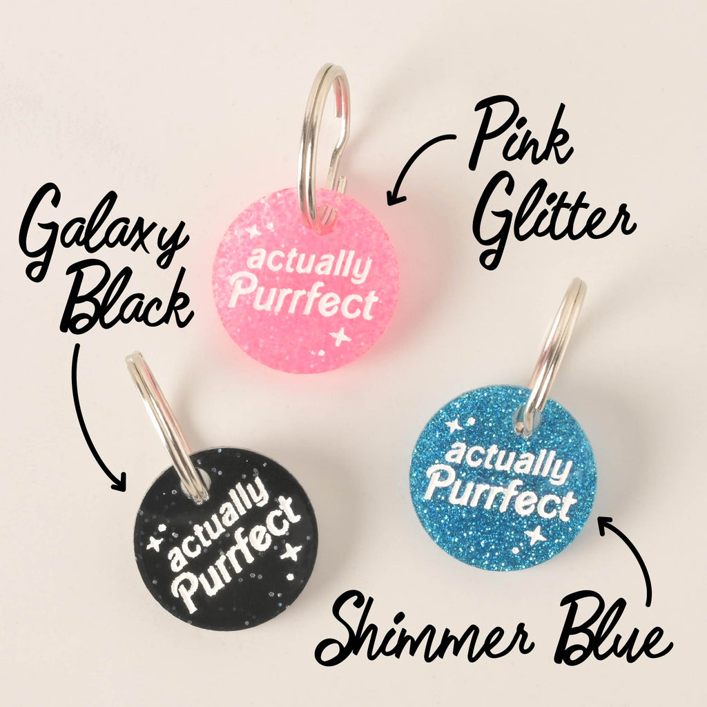 A product photo showing three round, glittery pet ID tags. The tag on the top is pink glitter, the tag on the bottom left is black glitter labeled "Galaxy Black," and the tag on the bottom right is blue glitter labeled "Shimmer Blue." All three tags are engraved with the words "actually Purrfect" and are shown with a silver split key ring.