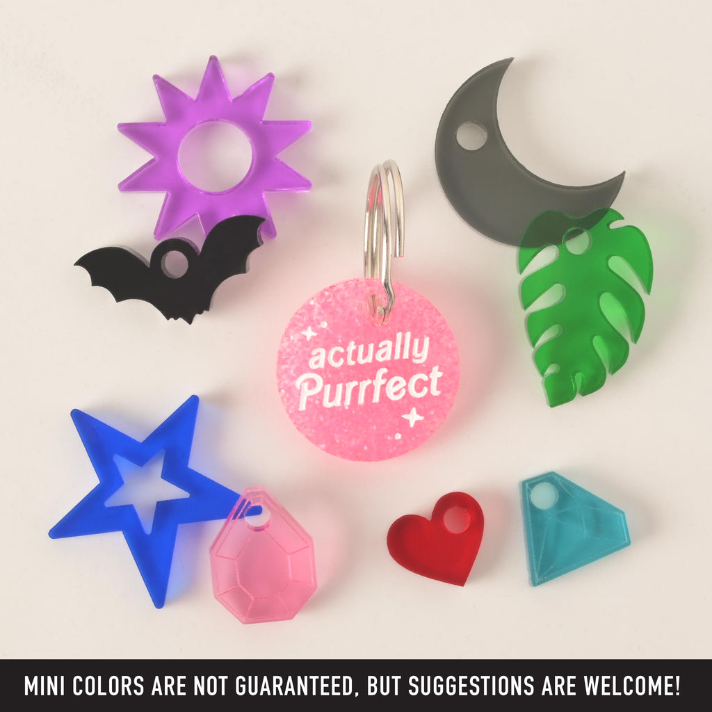 A product photo of a small, round, pink glittery tag with the words "actually Purrfect" in white text. Surrounding the main tag are several small, colorful charms: a purple sun, a black bat, a gray crescent moon, a green monstera leaf, a light blue geometric gem, a red heart, a pink faceted gem, and a blue star. Text at the bottom of the image reads, "MINI COLORS ARE NOT GUARANTEED, BUT SUGGESTIONS ARE WELCOME!"