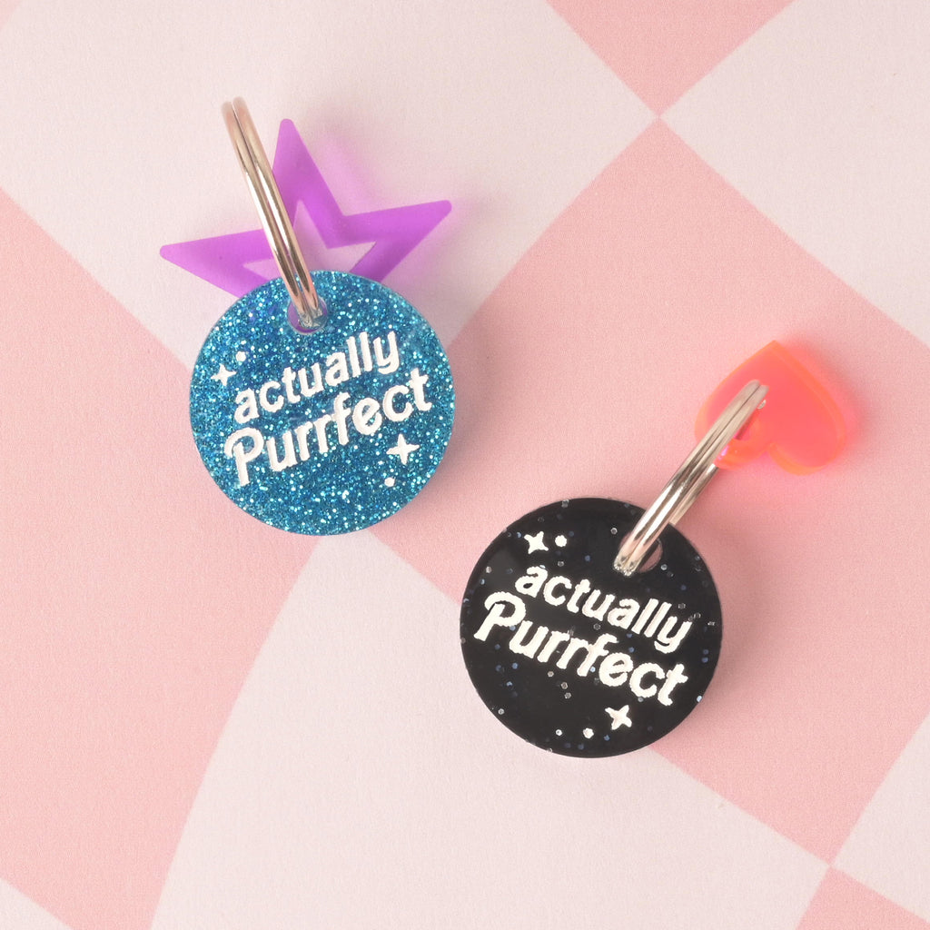 A pair of round pet tags, both featuring the text "actually Purrfect" in white letters. One tag is a blue glitter with a purple star charm attached, and the other is a black glitter with a pink heart charm. Both tags are secured with a silver ring and are displayed on a pink and white checkered background.