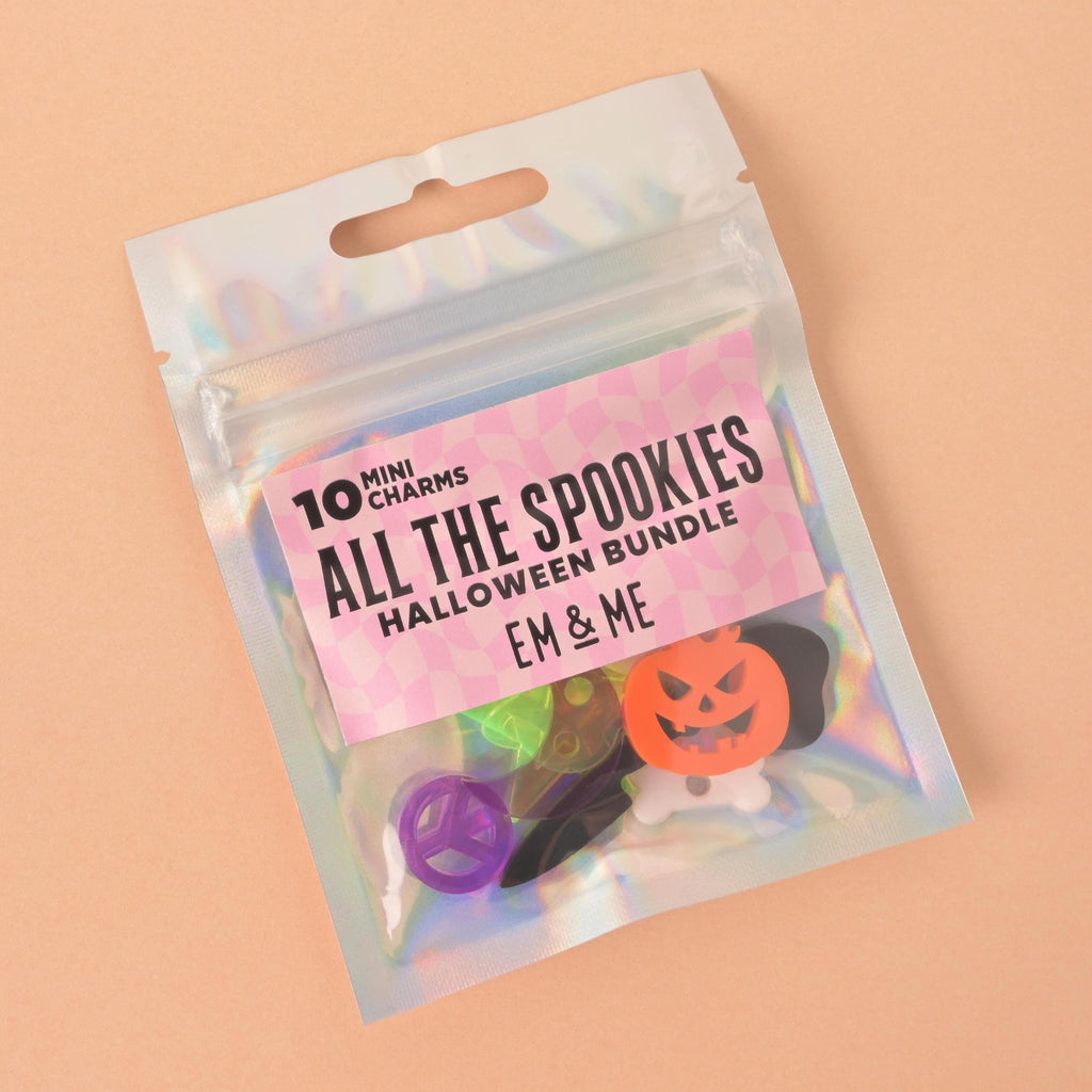 A clear, holographic bag containing the "All The Spookies" Halloween charm bundle from the brand "EM & ME." The bag is labeled "10 MINI CHARMS." Visible charms inside the bag include an orange jack-o'-lantern, a white bone, a purple peace sign, a black bat, and a translucent green star. The bag is shown on a solid peach-colored background.