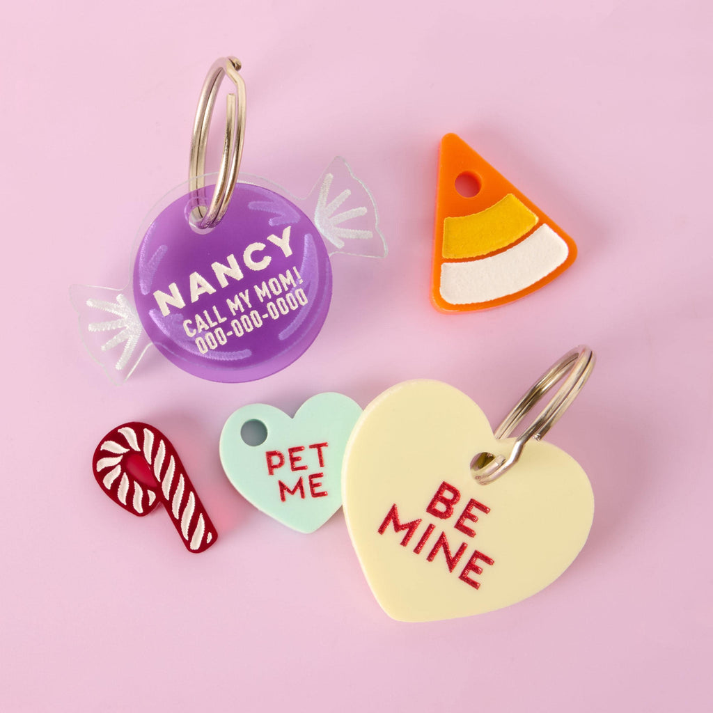 A collection of assorted pet tags and charms in candy shapes scattered on a solid pink background. The items include a personalized purple candy wrapper tag for "NANCY," a yellow conversation heart tag that says "BE MINE," a mint green heart charm that says "PET ME," a candy corn charm, and a small candy cane charm.