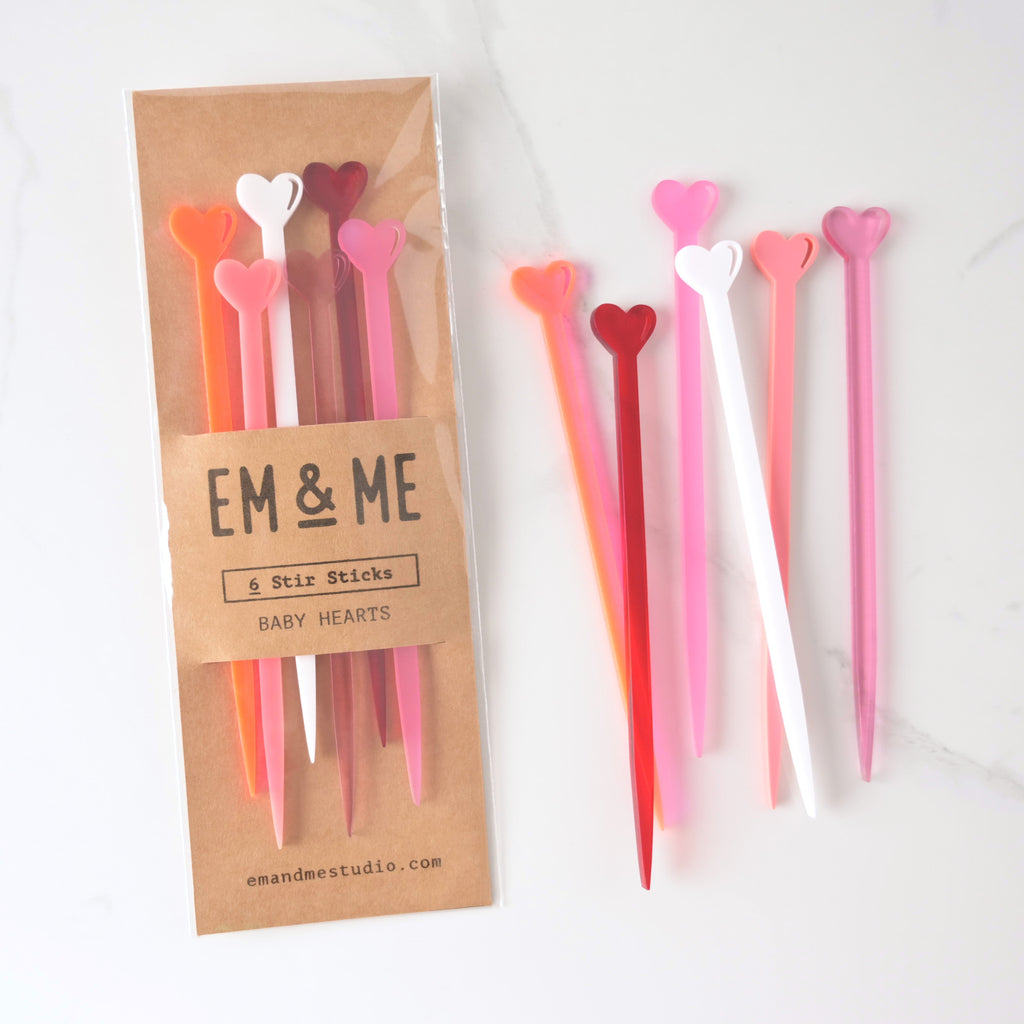 Collection of tiny heart-shaped cocktail stirrers in various shades of pink, red and white packaged on brown Kraft paper with more stir sticks off to the side on marble background. Fluorescent pink, red, white, frosted pink, opaque pink and transparent pale pink