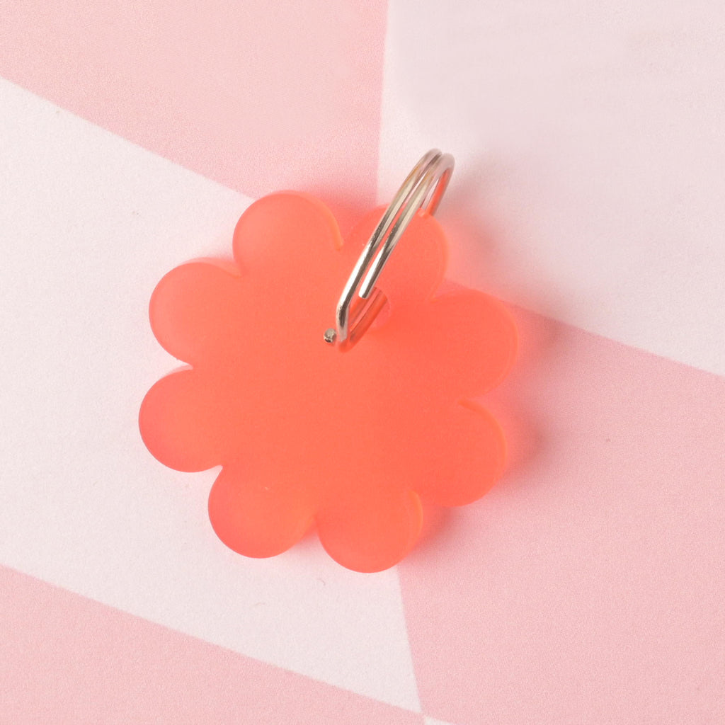 Rear view of a bright red-orange flower-shaped pet ID tag called "Poppy" with a metal keyring attached, placed on a pink checkered background