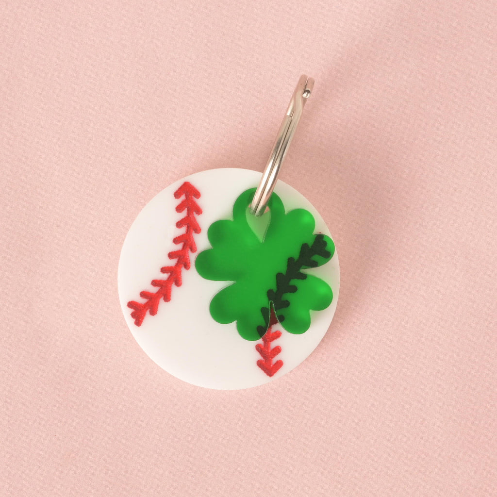 A custom, white, circular pet ID tag with a red engraved baseball stitch pattern. A separate, translucent green, four-leaf clover charm with a black and red baseball stitch pattern is attached to the silver keyring. Both charms are displayed on a solid pink background.