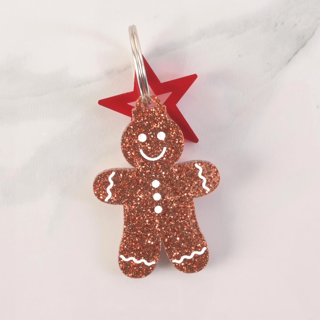 A close-up of a glittery copper gingerbread man keychain or pet tag, with a smiling face and white icing details. A red star charm is attached behind the gingerbread man. Both are connected to a silver ring and are displayed on a white marble surface.