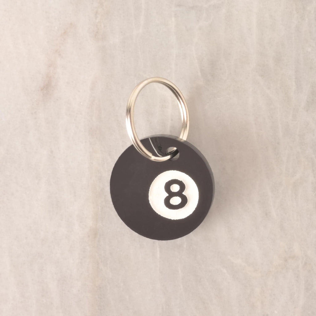 Round black acrylic pet ID tag styled like an 8-ball, with a metal keyring, photographed on a neutral stone background.