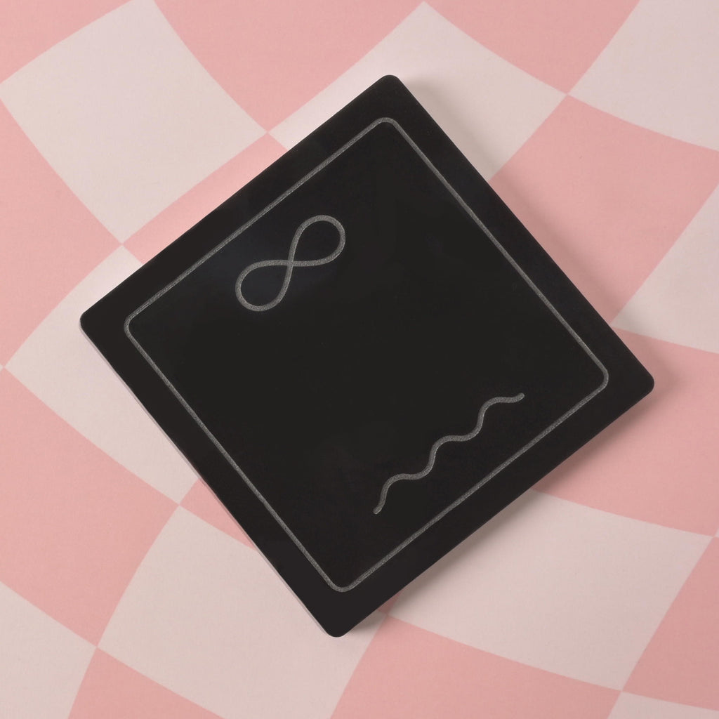 A single black, square-shaped coaster is shown on a pink and white checkered background. It has a silver-etched design of an infinity symbol and a wavy line.