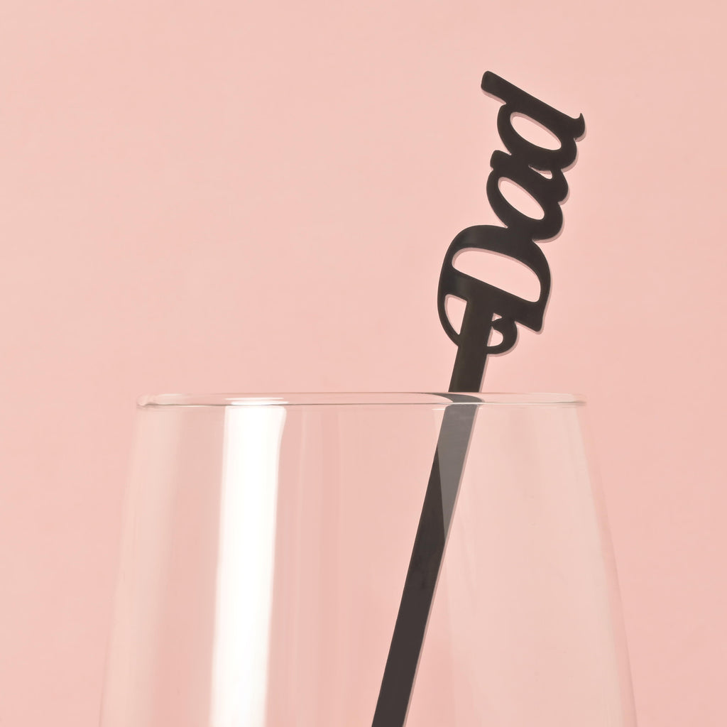 A single black cocktail stirrer with the word "Dad" is placed inside a clear glass. The background is a solid pink color.