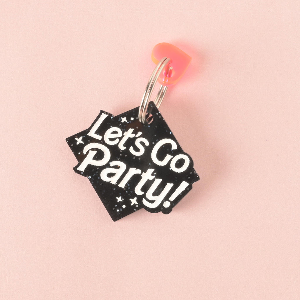 A close-up product photo of a black glittery, rhombus-shaped pet tag with the words "Let's Go Party!" engraved on it. A neon orange heart charm is attached to the top of the tag, which is shown with a silver split key ring.