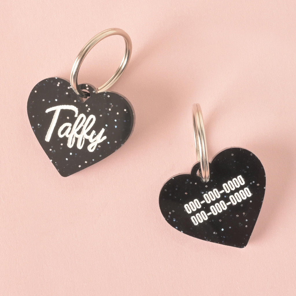A product photo showing two sides of the same black glittery, heart-shaped pet tag. The tag on the left shows the front with the name "Taffy." The tag on the right shows the back, which is custom engraved with two lines for phone numbers. Both tags have a silver split key ring attached.