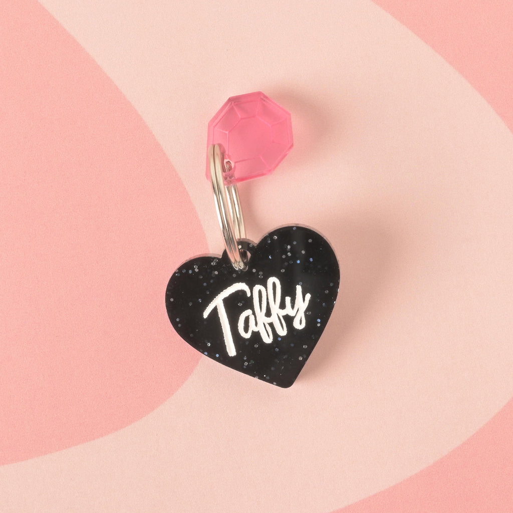 A close-up product photo of a black glittery, heart-shaped pet tag with the name "Taffy" engraved on it. A translucent pink gemstone charm is attached to the top of the tag, which is shown with a silver split key ring.