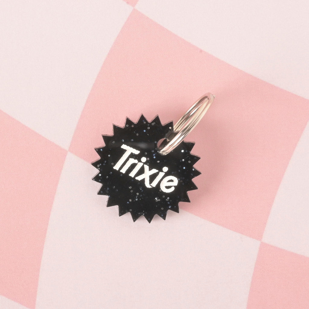 A close-up product photo of a black glittery pet ID tag with a unique starburst or spiked edge. The tag is engraved with the name "Trixie" and is shown with a silver split key ring on a checkered pink and white background.