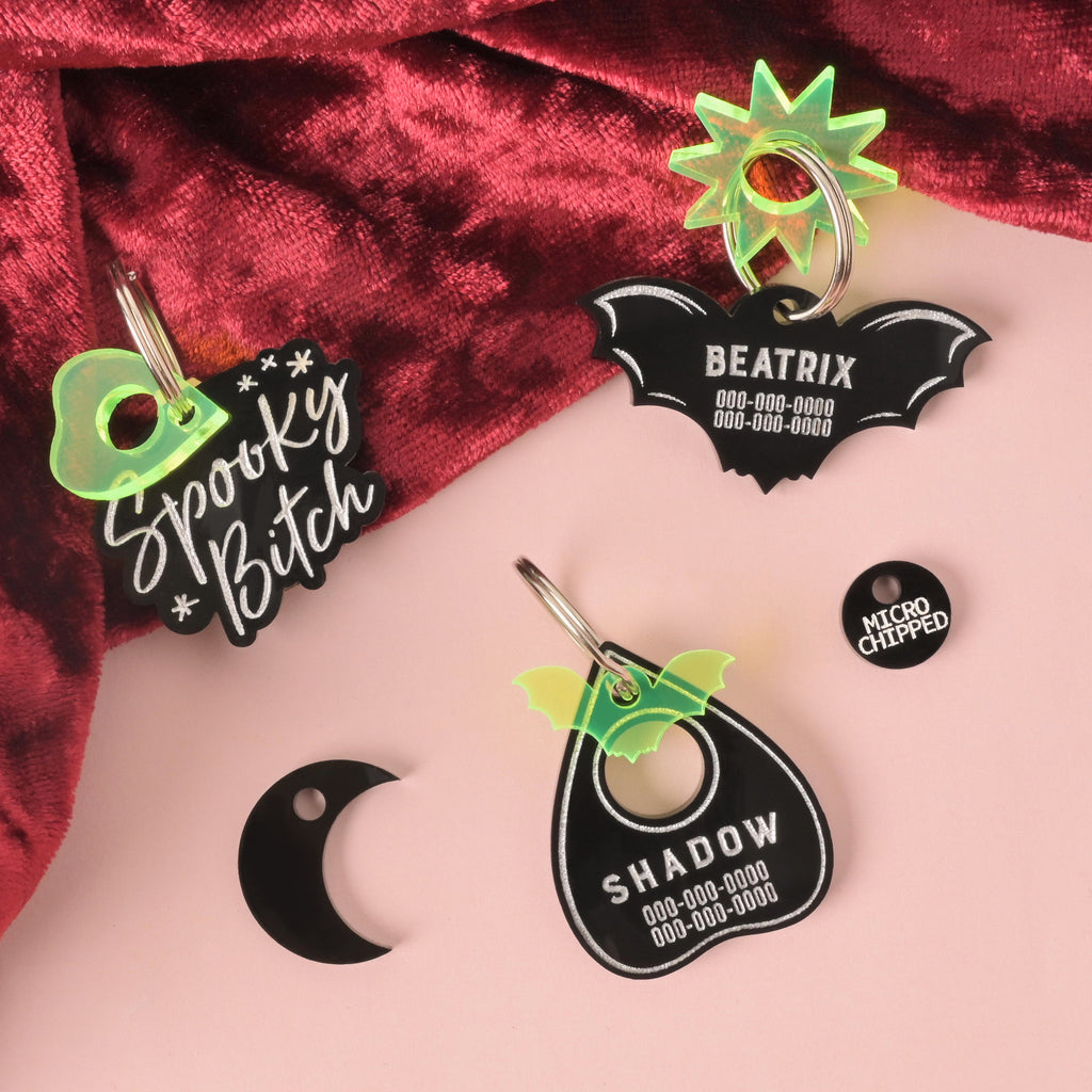 Collection of Black, bat-shaped, Ouija planchet-shaped and "spooky bitch" pet tags with fluorescent green mini charms on a solid pink background with red velvet.