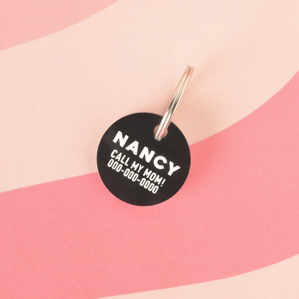 A close-up product photo of a round, black pet tag, also known as a Minimal Circle tag. The tag is engraved with the name "NANCY" and the funny phrase "CALL MY MOM!" in white text. It is shown with a silver split key ring.