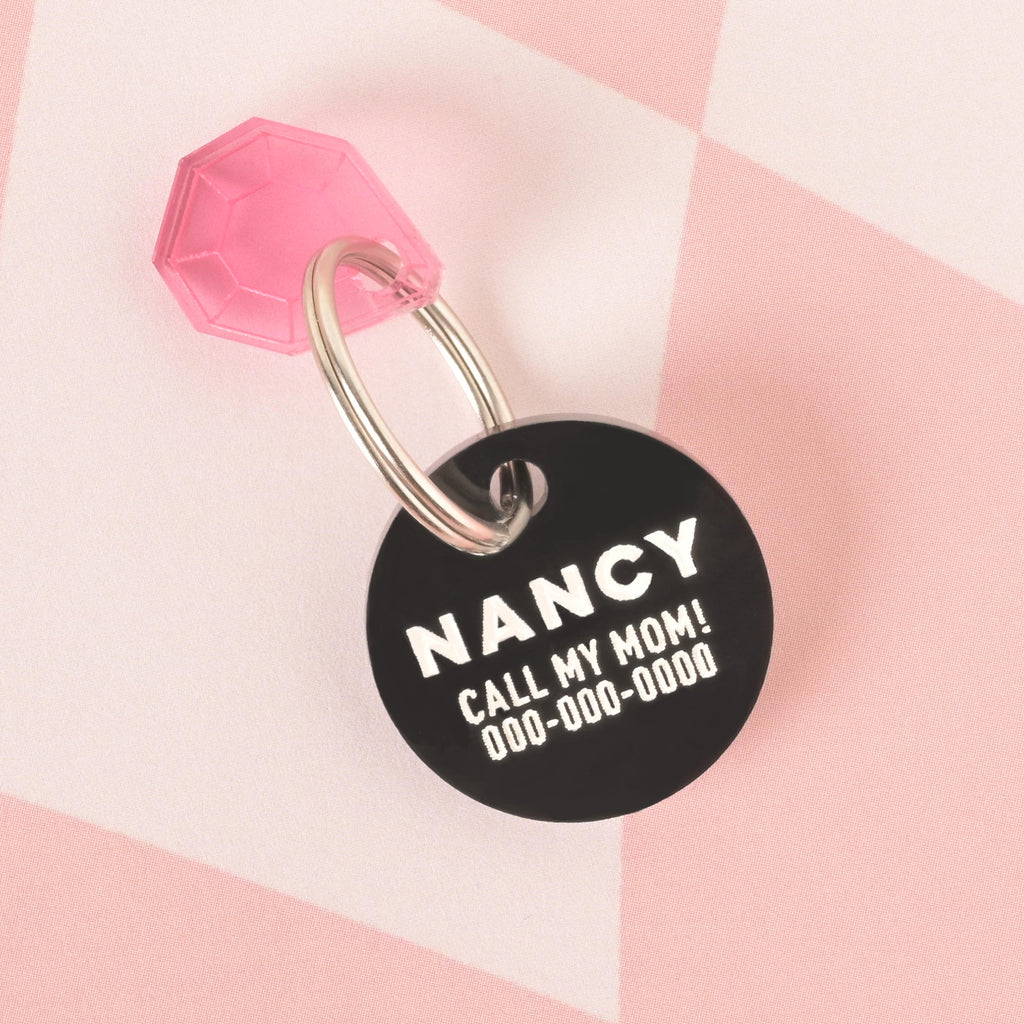 A close-up product photo of a black, round pet ID tag, also known as a Minimal Circle tag. The tag is engraved with the name "NANCY" and the funny phrase "CALL MY MOM!" in white text. A translucent pink gemstone charm is attached to the top of the tag, which is shown with a silver split key ring.