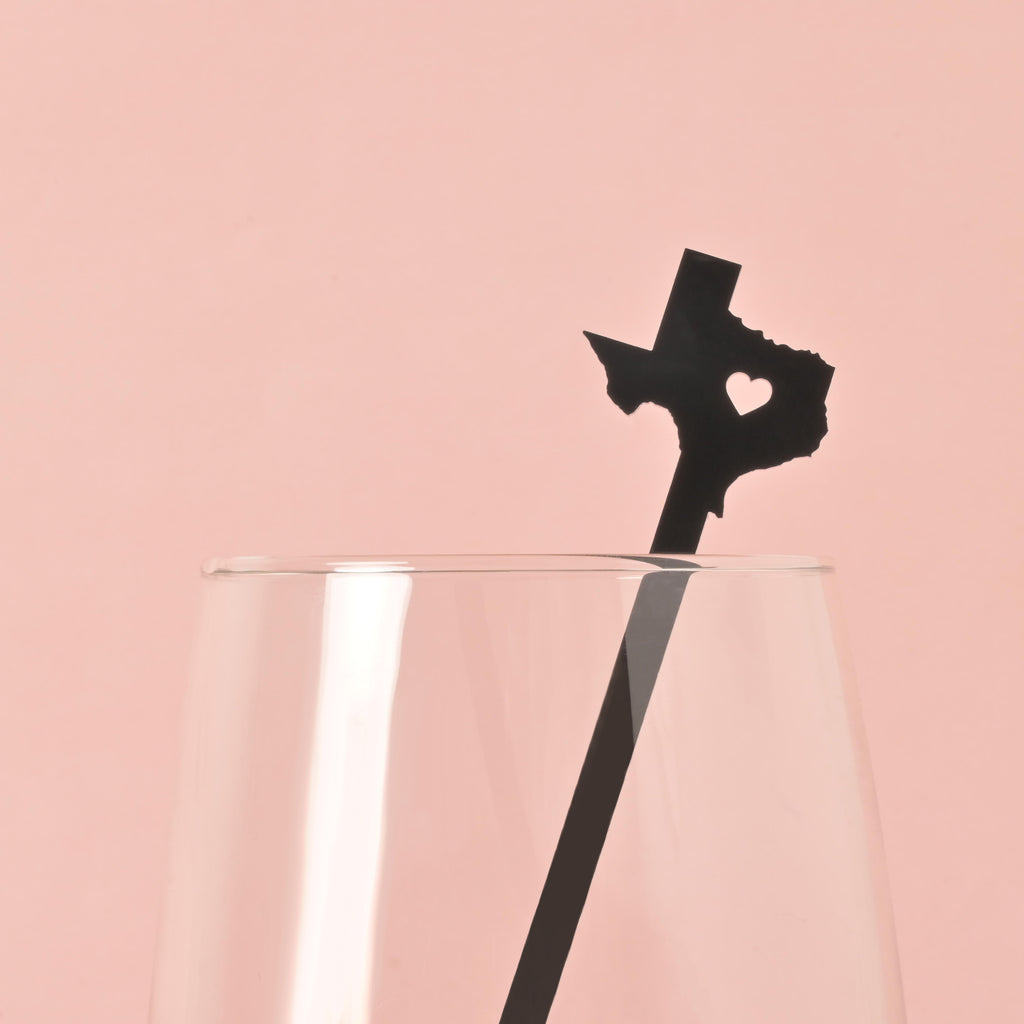 A single black cocktail stirrer shaped like the state of Texas with a heart cut out in the middle is placed inside a clear glass. The background is a solid pink color.