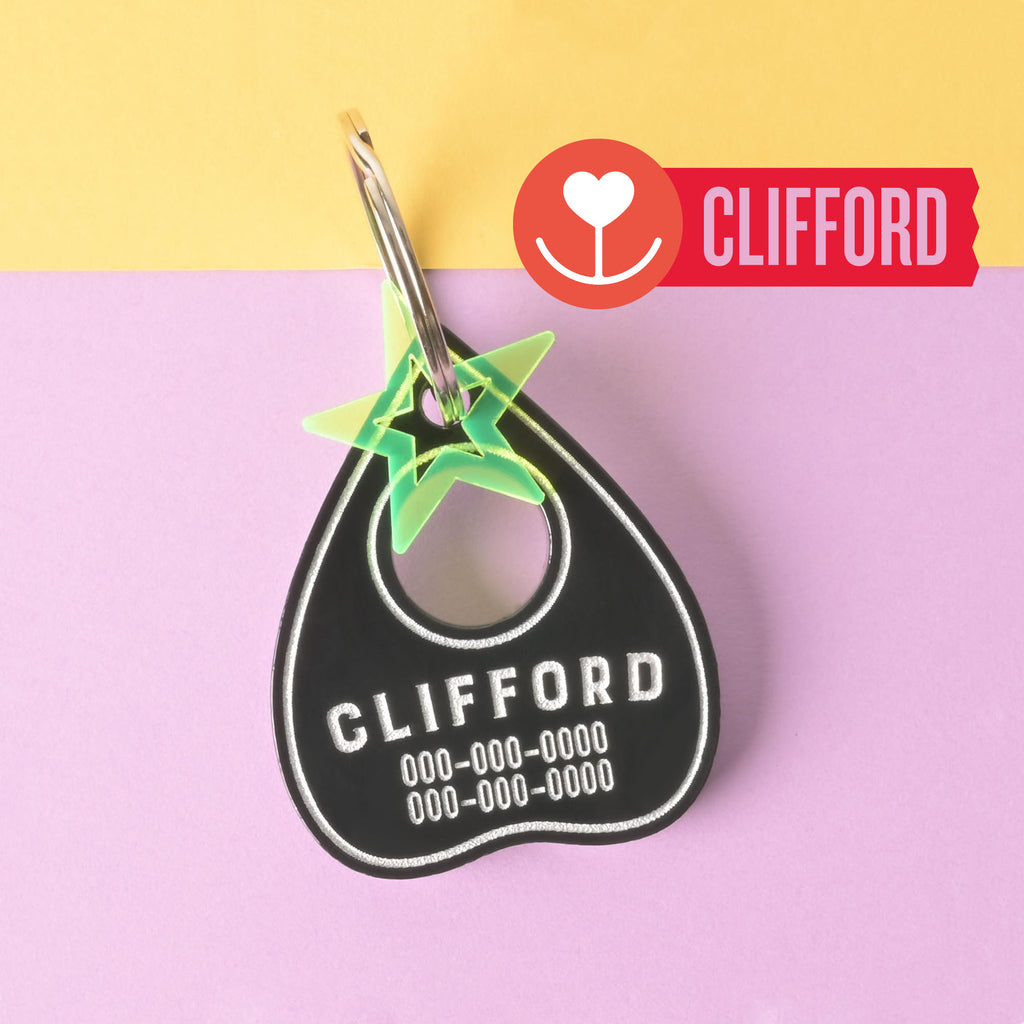 A black planchette-shaped pet ID tag for Clifford, showing contact phone numbers, attached to a metal snap clip and a neon green star charm. The tag is displayed against a yellow and lavender background.