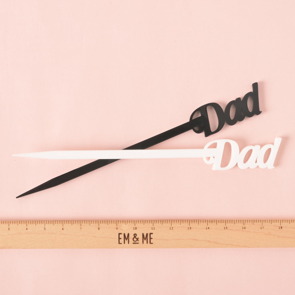 A pair of "Dad" cocktail stirrers—one black and one white—are crossed over each other. A ruler with the "EM & ME" logo is placed below the stirrers to show their size. The background is a solid pink color.