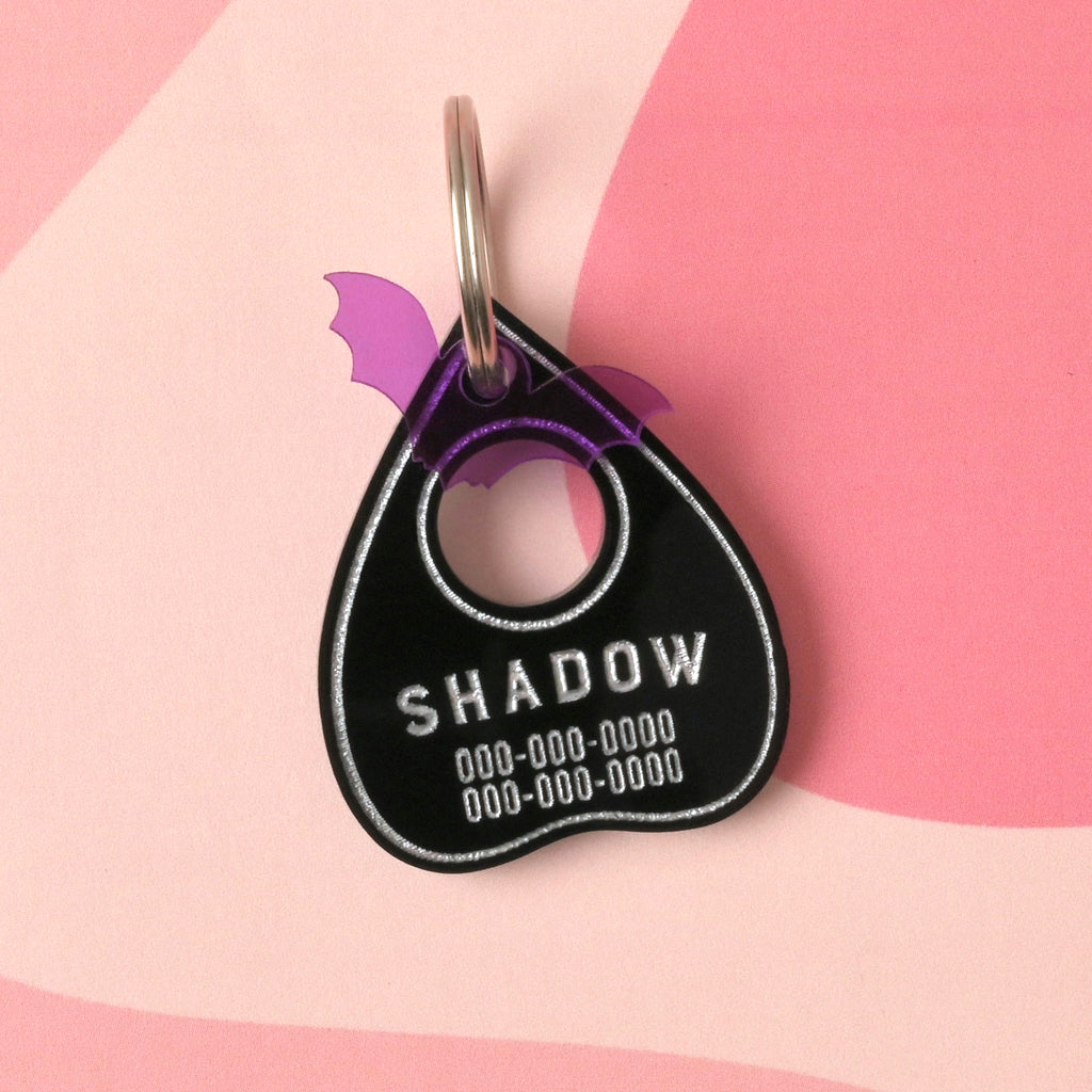 Opaque black ouija planchet0shaped pet tag with a purple bat mini charm on pink wavy background.