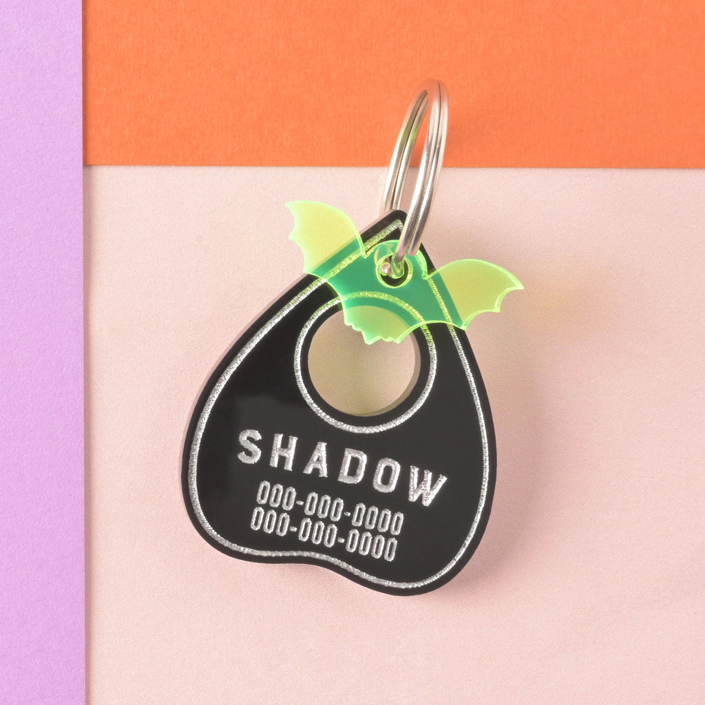 Black ouija planchet-shaped pet tag with fluorescent green bat mini charm on color blocked background