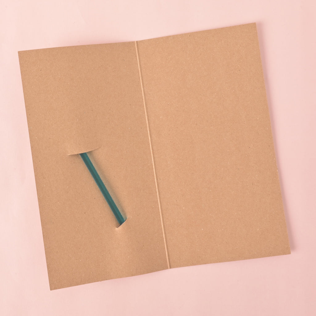 An Blank brown gift card, folded in half, with a slit cut into it to hold a stirrer. The back of a teal cocktail stirrer is partially visible through the slit. The background is a solid pink color.