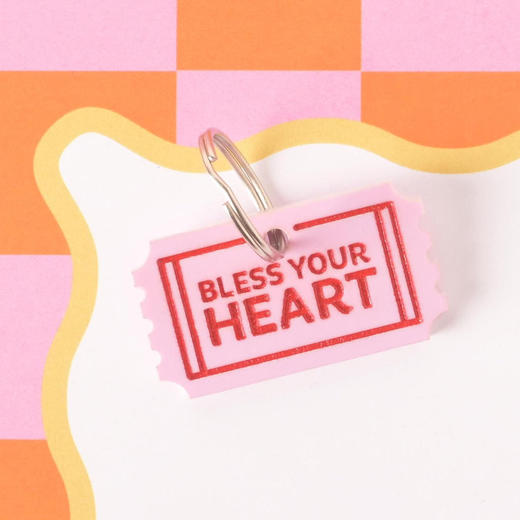 Pink acrylic pet charm shaped like a ticket with red Bless Your Heart text, attached to a ring for use as a playful pet collar accessory.