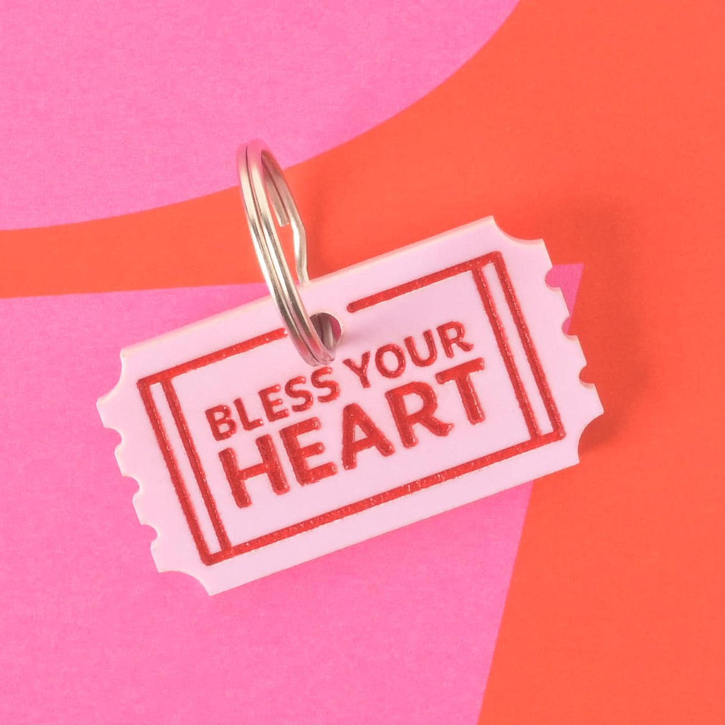 Pink acrylic ticket-shaped pet ID tag with red “Bless Your Heart” lettering, shown with silver keyring on a colorful pink and red background.