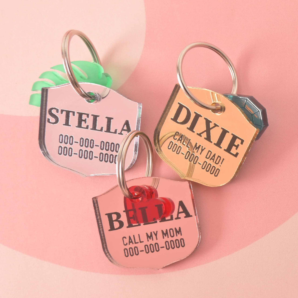 A set of three personalized pet tags in a shield shape. The tags include a pink one for "STELLA," a gold one for "DIXIE," and a red one for "BELLA." Each tag has a placeholder phone number and is attached to a silver ring for a collar. The items are displayed on a pink background with a wavy design.