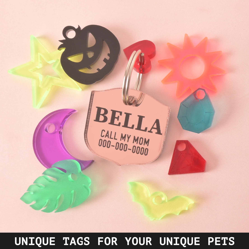 A personalized rose gold shield-shaped pet tag with the name "BELLA" and a placeholder phone number. Surrounding the tag are various charms, including a black carved pumpkin, a neon yellow star, an orange sunburst, a purple moon, a green monstera leaf, and a translucent yellow bat. There are also red and blue diamond-shaped charms. The image contains the text "UNIQUE TAGS FOR YOUR UNIQUE PETS." The items are displayed on a solid pink background.