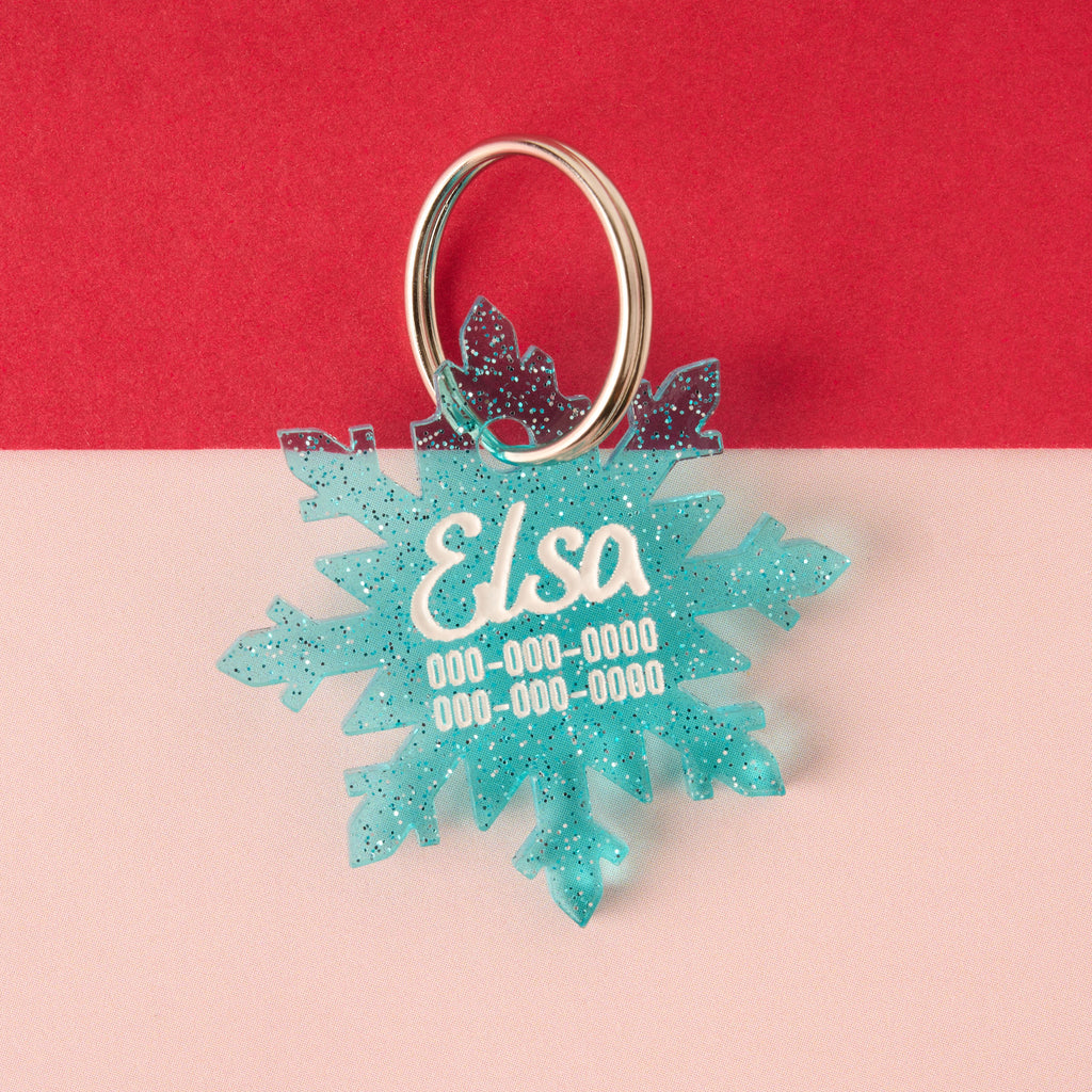 A custom, laser-cut pet ID tag shaped like a large snowflake in glittery blue acrylic. The tag is engraved with the name 'Elsa' and three lines of contact information in white, set against a red and white background.