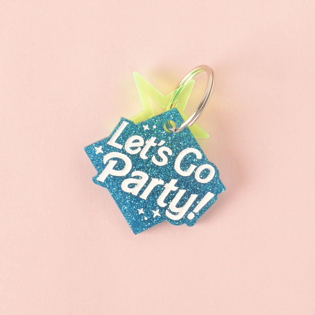 A close-up product photo of a blue glittery, rhombus-shaped pet tag with the words "Let's Go Party!" engraved on it. A neon yellow star charm is attached to the top of the tag, which is shown with a silver split key ring.