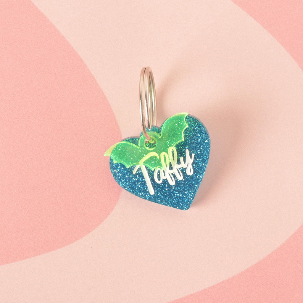 A close-up product photo of a blue glittery, heart-shaped pet tag with the name "Taffy" engraved on it. A neon green glitter bat wings charm is attached to the top of the tag, which is shown with a silver split key ring.