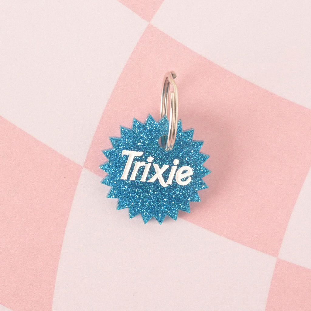 A close-up product photo of a blue glittery pet ID tag with a unique starburst or spiked edge. The tag is engraved with the name "Trixie" and is shown with a silver split key ring on a checkered pink and white background.