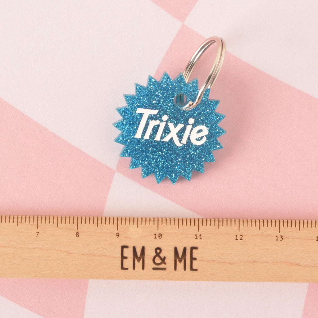 A close-up product photo of a blue glittery pet ID tag with a unique starburst or spiked edge. The tag is engraved with the name "Trixie" and is shown with a silver split key ring. A wooden ruler with the words "EM & ME" is positioned below the tag to show its size.