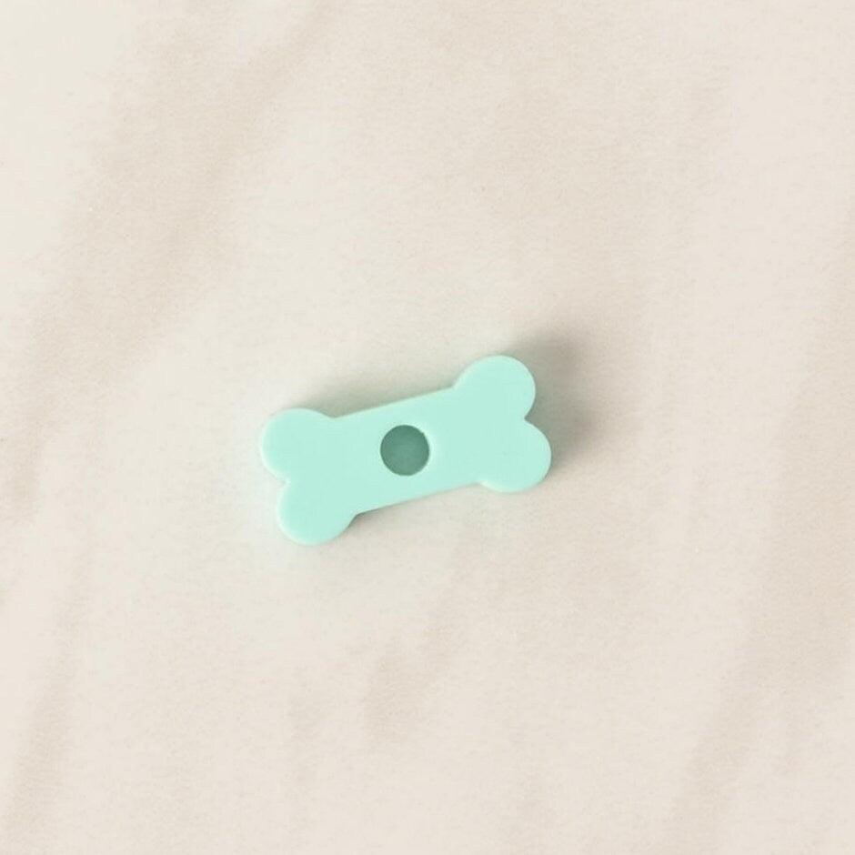 A close-up of a small, translucent mint green, bone-shaped pet tag charm. The charm is displayed on a light-colored, speckled countertop or stone surface.