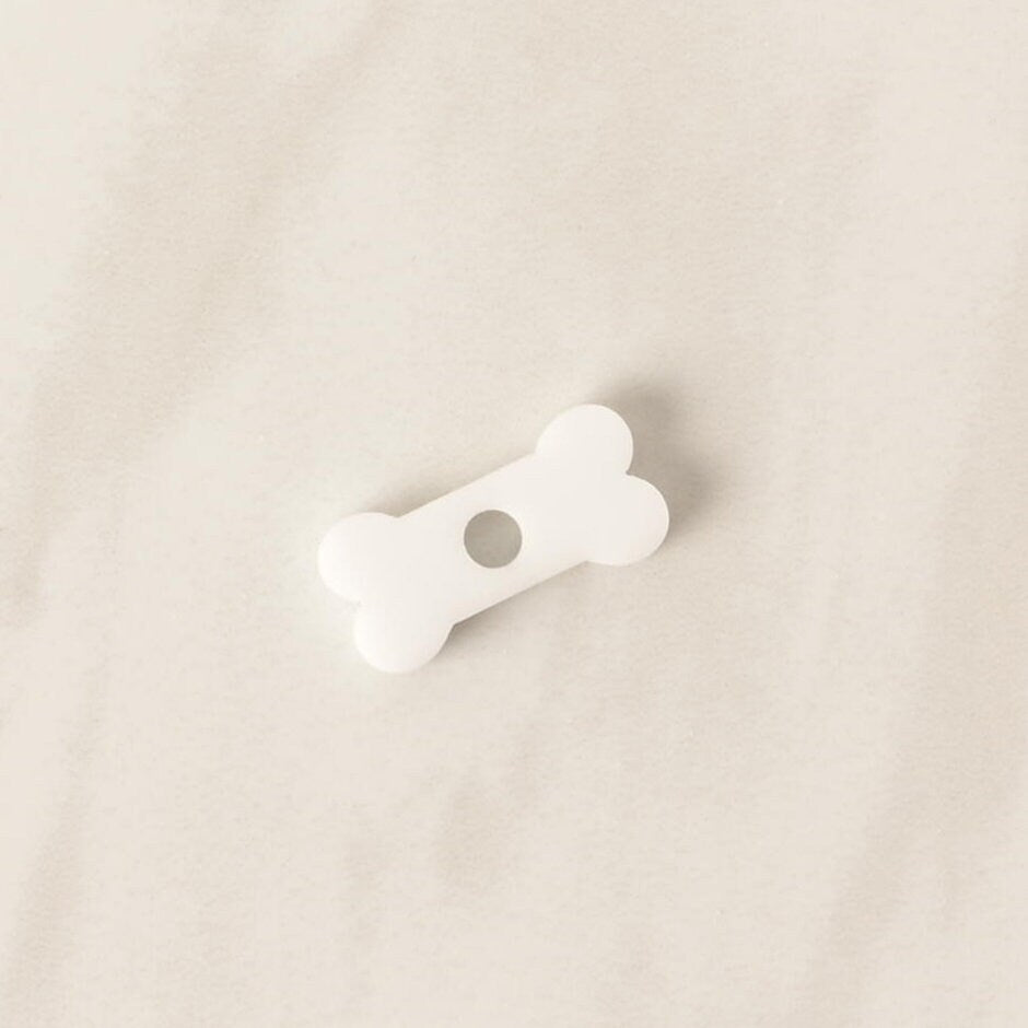 A close-up of a small, white, bone-shaped pet tag charm. The charm is displayed on a light-colored, speckled countertop or stone surface.
