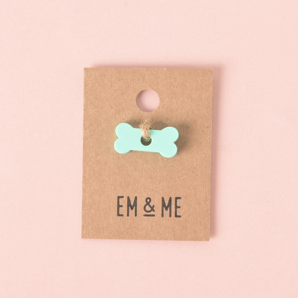 A small, bone-shaped pet tag charm in a pastel mint green color. The charm is attached with a piece of string to a brown cardboard tag with the logo "EM & ME." The item is displayed on a solid pink background.