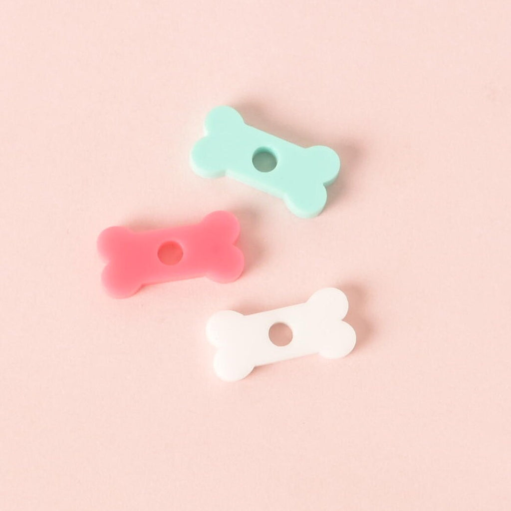 A group of three small, bone-shaped pet tag charms in pastel colors: white, light pink, and opaque teal. The charms are arranged on a solid pink background.