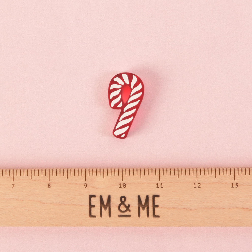 A red and white candy cane-shaped charm. A wooden ruler with the "EM & ME" logo is placed below the charm to show its size. The charm is shown on a solid pink background.