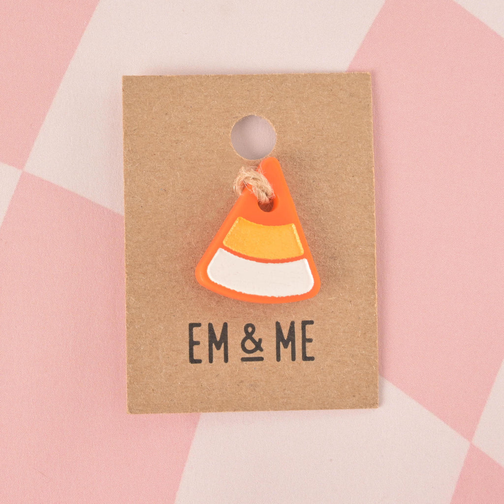 A single candy corn-shaped pet tag charm. The charm is orange, white, and yellow and is attached to a piece of string. It is displayed on a small brown cardboard tag with the logo "EM & ME" printed on it. The background is a pink and white checkered pattern.